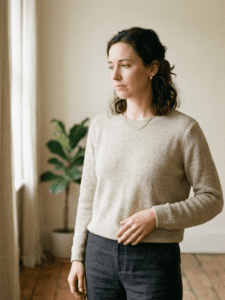 A woman standing in soft natural light near a window, one hand resting lightly on her side, looking thoughtful. Clean, neutral background. Calm but slightly “off” expression.